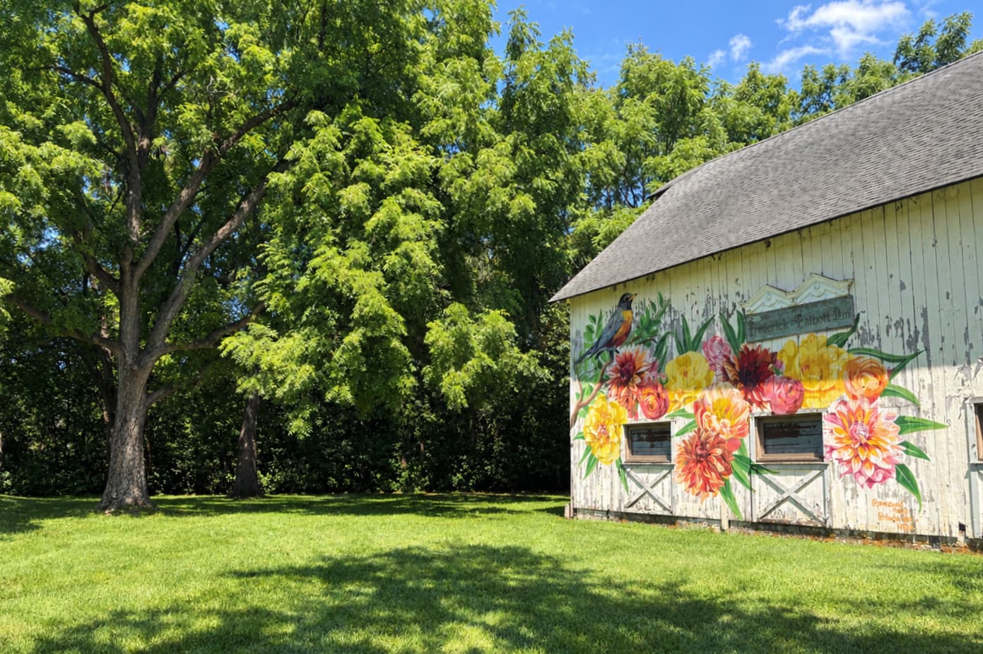 A vibrant mural of flowers adorns a rustic barn beside a lush green lawn and a large tree.