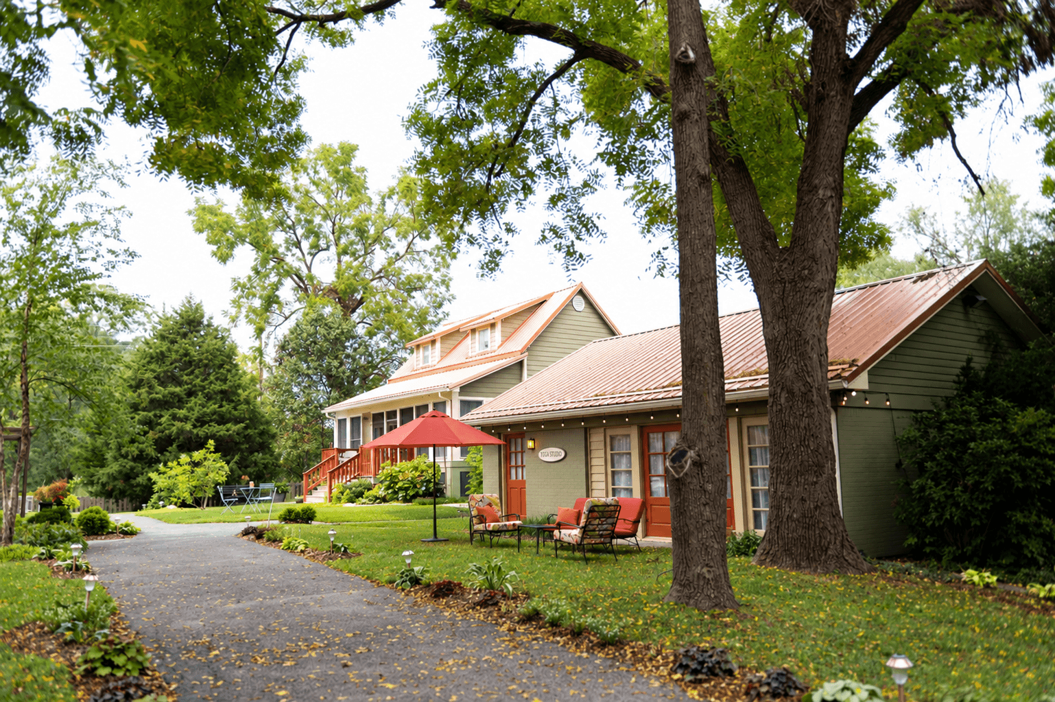 A charming pathway leads to two rustic houses surrounded by green trees and landscaping.