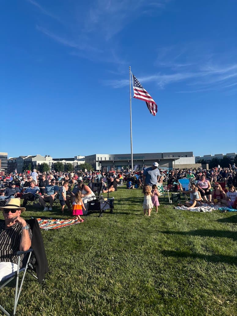 A large crowd gathers on a sunny day in a park, with an American flag flying prominently.