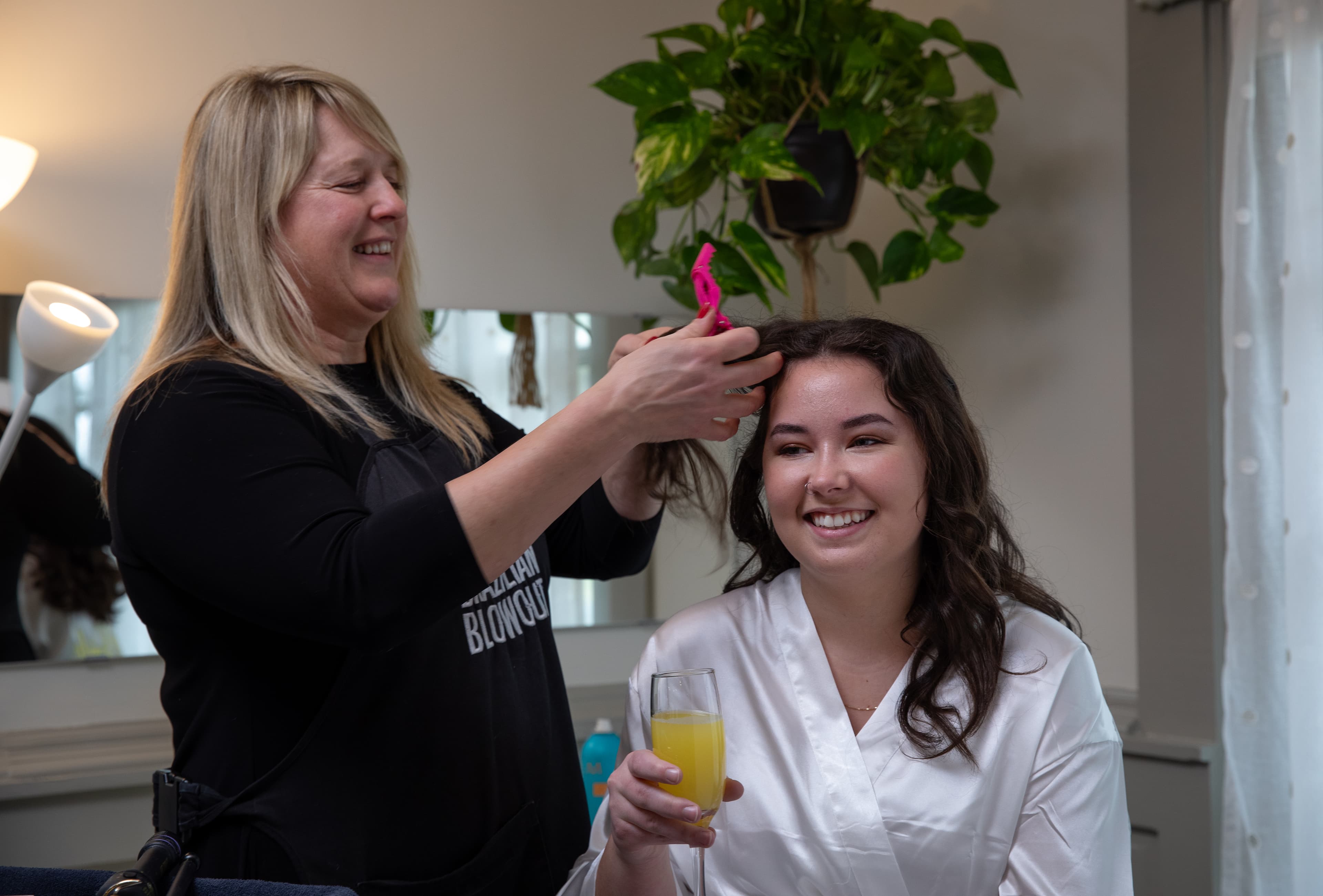 A hairstylist smiles while curling the hair of a woman in a white robe, who holds a glass of orange juice.