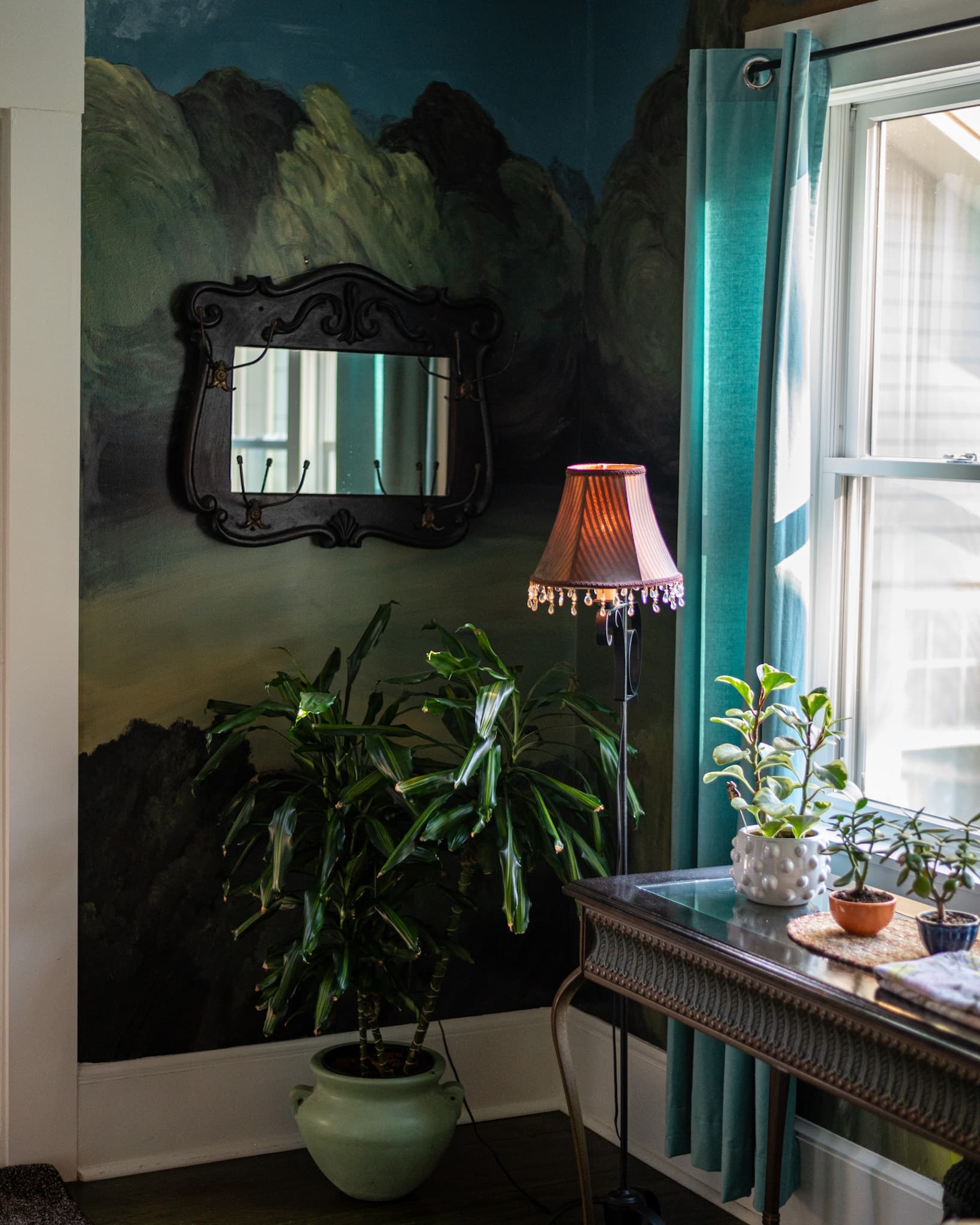 A cozy corner featuring a vintage lamp, potted plants, a decorative mirror, and teal curtains against a nature-themed mural.