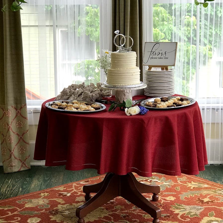 A round table dressed in red holds a wedding cake, assorted desserts, and a sign saying "favors please take one."