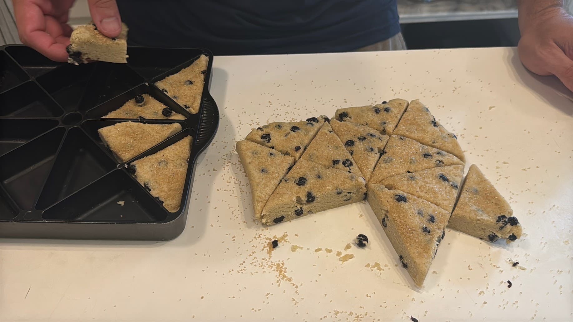 A person is removing a piece of unbaked scone dough with dark berries from a tray on a countertop.