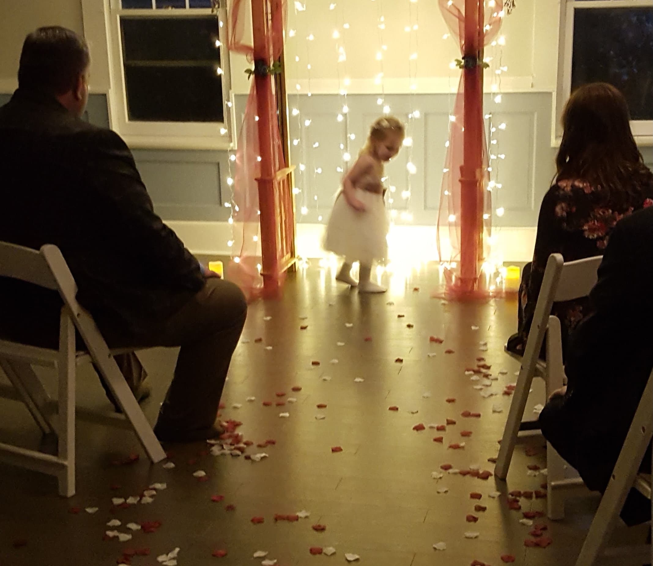 A young girl in a white dress walks down an aisle decorated with rose petals, flanked by guests seated in chairs.