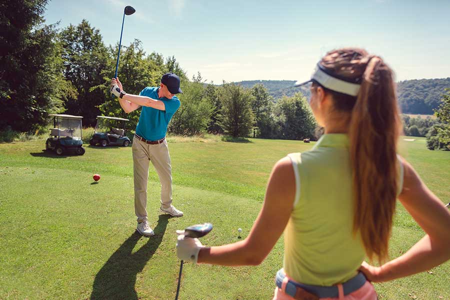A golfer swings a club while a woman watches on a sunny golf course.