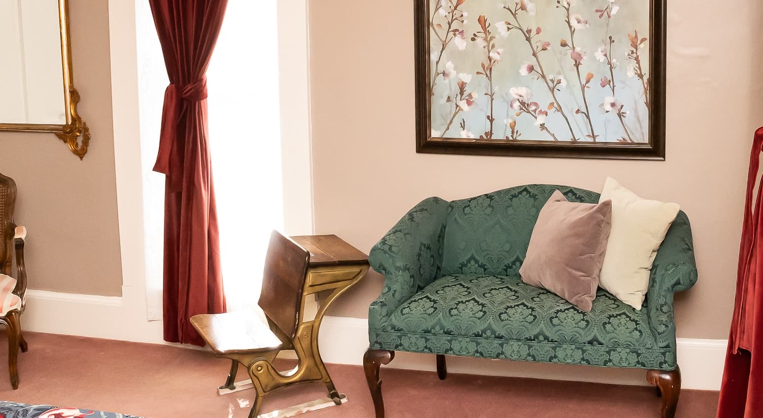 A teal upholstered sofa with decorative pillows sits beside a vintage brass chair in a softly lit room with a floral painting on the wall.