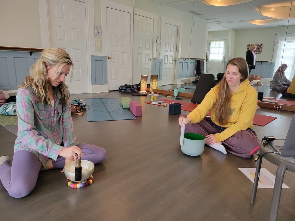 Two women are seated on the floor in a yoga studio, one playing a singing bowl while the other prepares to strike a green bowl.