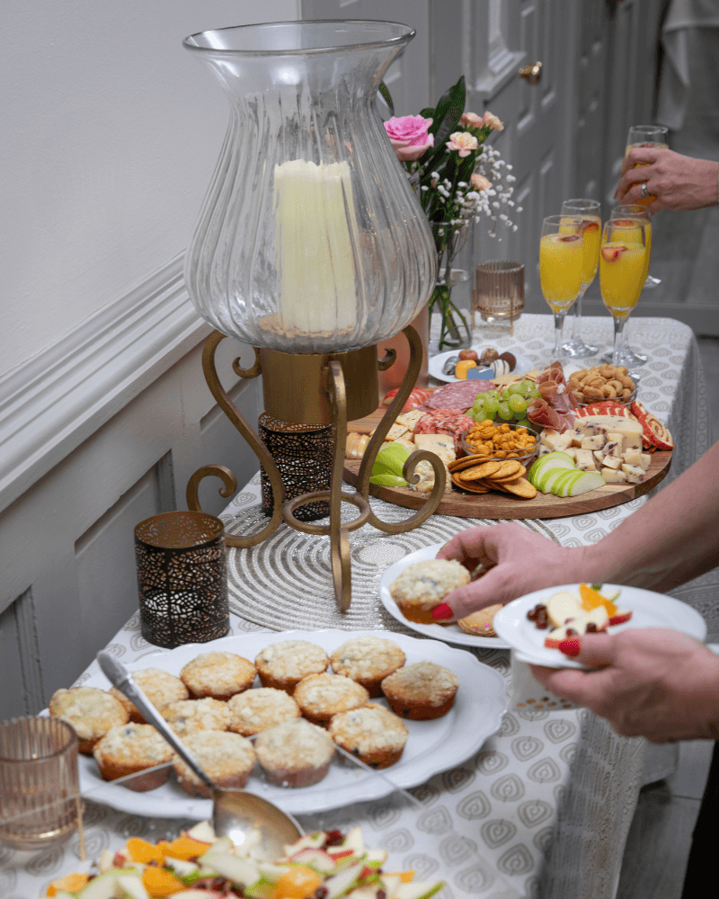 A table is set with an assortment of muffins, fruit, snacks, cheeses, and beverages, with a hand reaching for food.