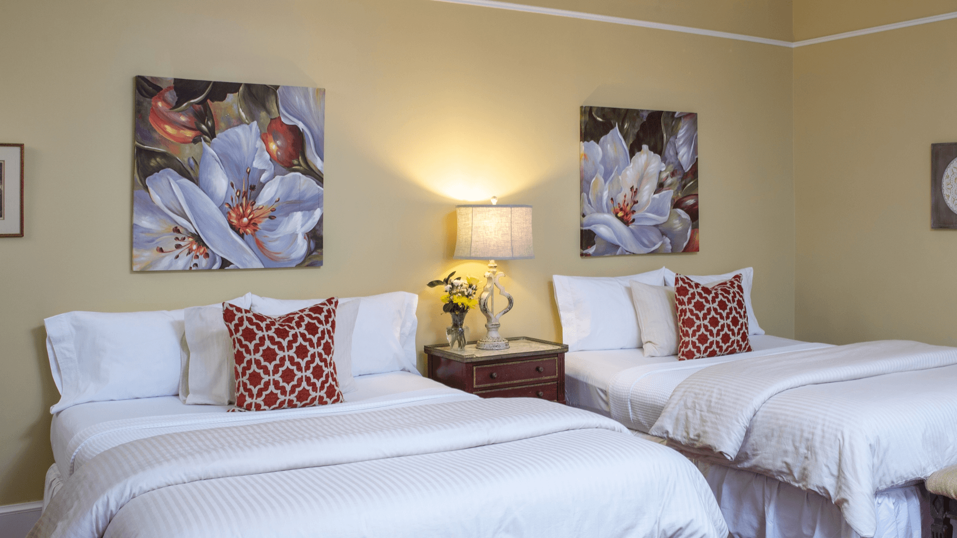 A cozy guest room featuring two beds, floral artwork, and a bedside lamp.