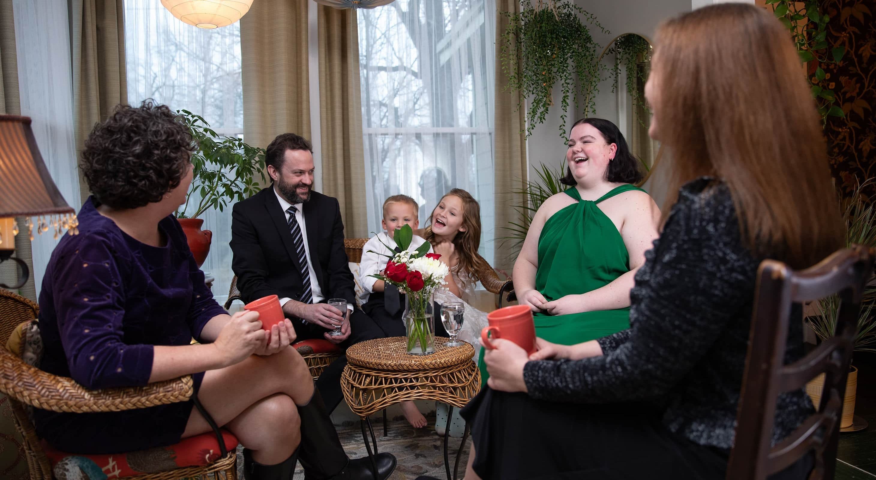A group of five adults and two children share smiles and conversation in a cozy, well-lit living room.