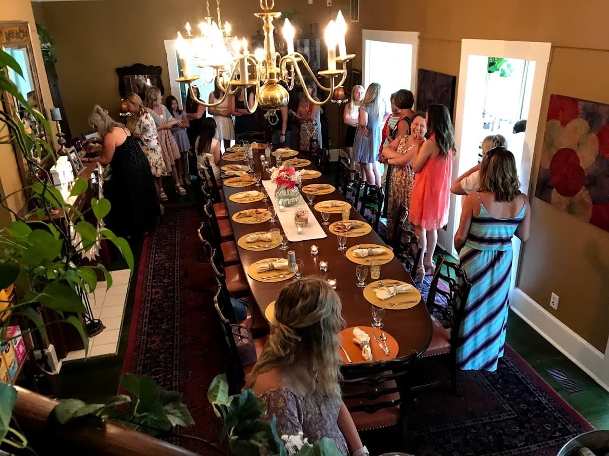 A group of women gather around a long dining table adorned with flowers and dishes in an elegantly decorated room.