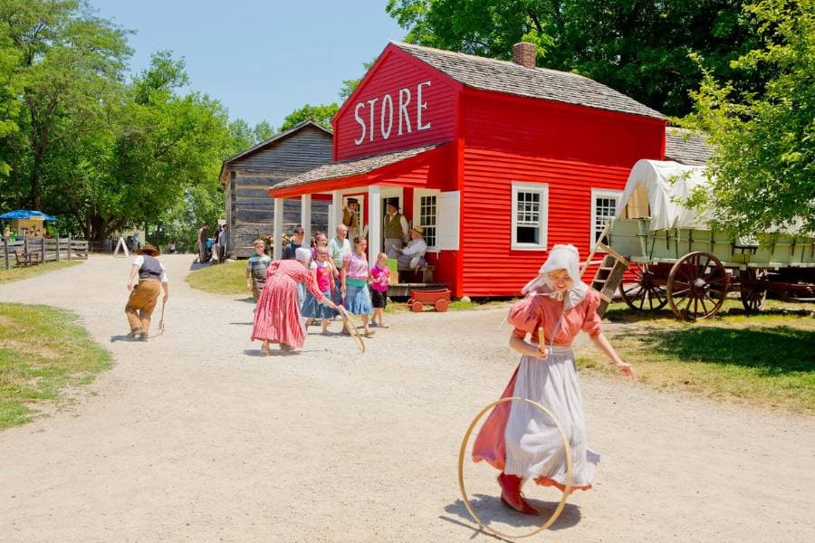 A historic village scene featuring a bright red store and people in period clothing engaging in various activities. A historic village scene featuring a bright red store and people in period clothing engaging in various activities.
