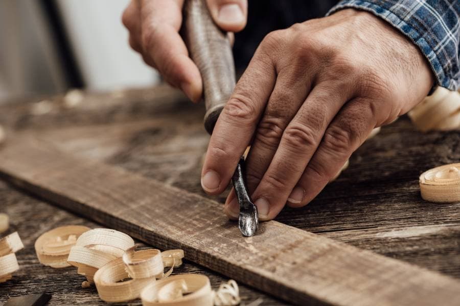 A person carves wood using a chisel, with shavings scattered on a wooden surface.
