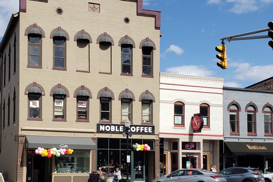 A street view of buildings featuring a coffee shop called "Noble Coffee," colorful balloon decorations, and a variety of storefronts.