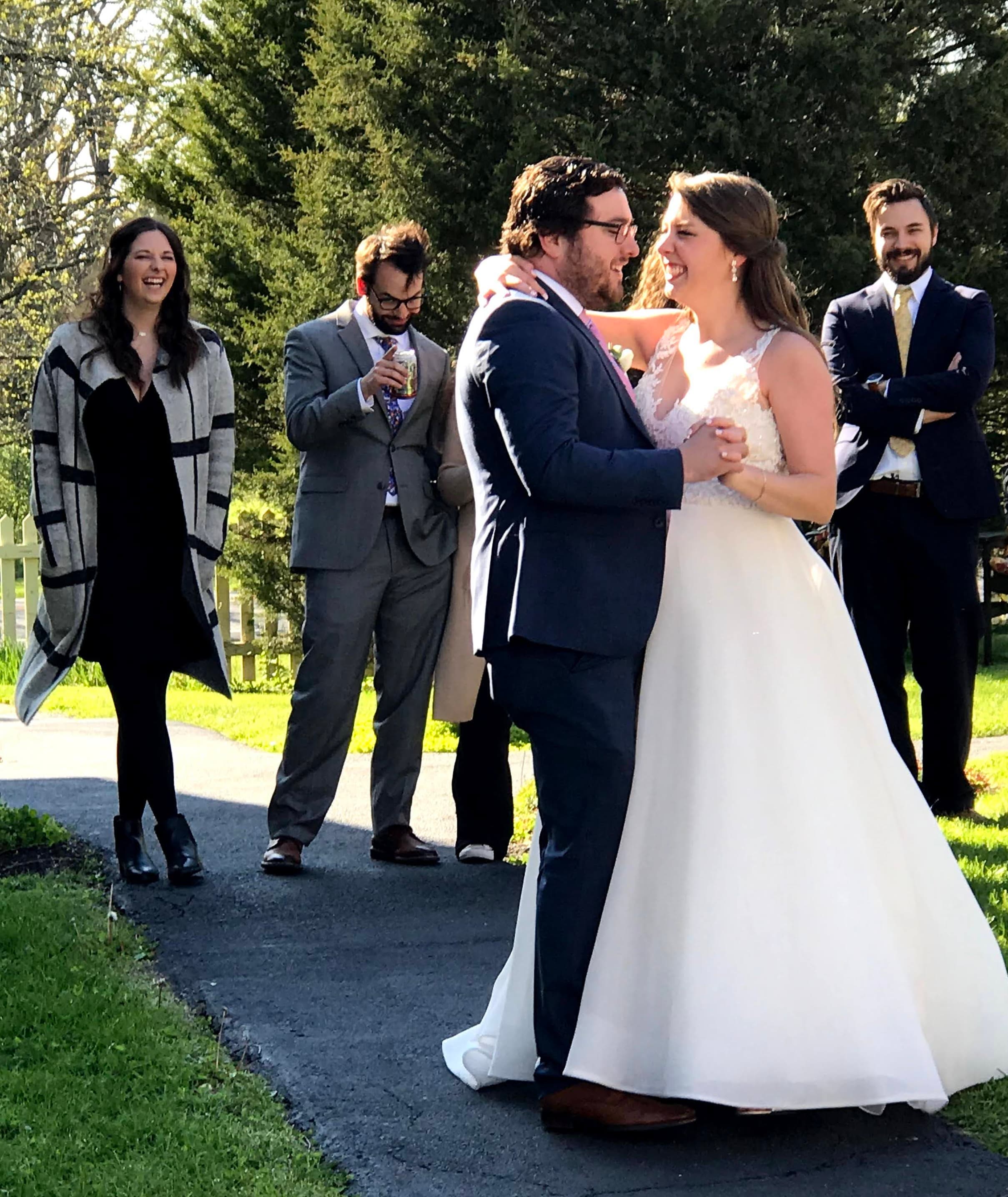 A bride and groom dance together joyfully while guests watch and smile in a lush outdoor setting.