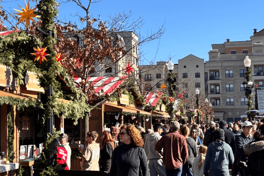 A busy outdoor market with festive decorations and crowds enjoying the day under a clear blue sky.