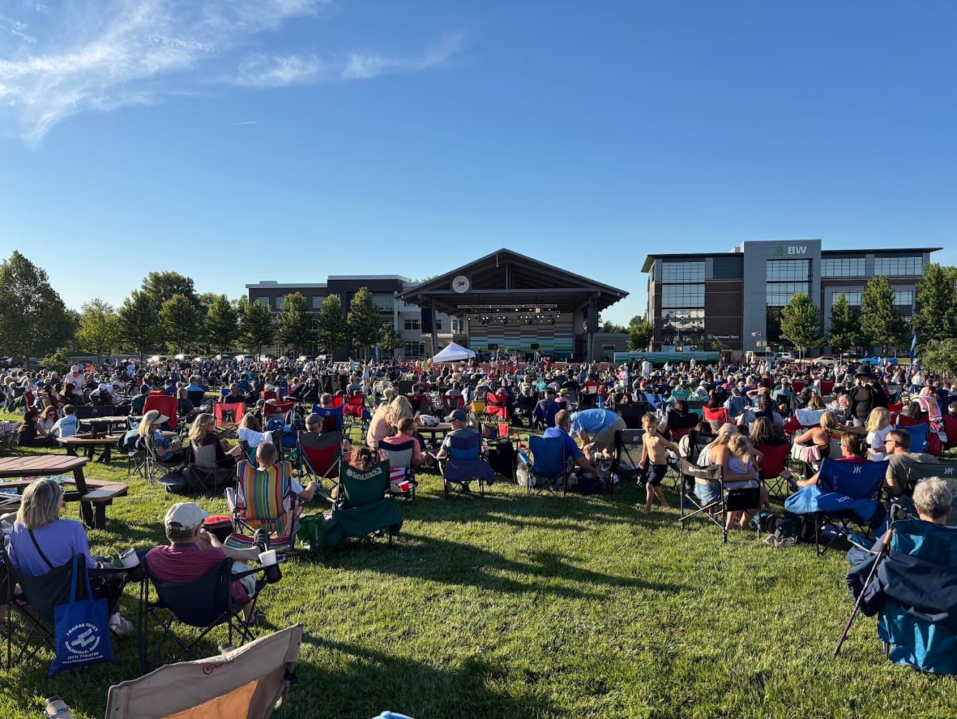 A large crowd gathers on a lawn in front of an outdoor stage during a sunny event. A large crowd gathers on a lawn in front of an outdoor stage during a sunny event.