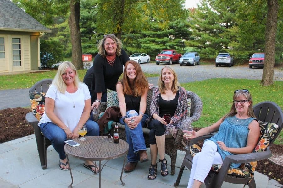 Five women sitting together on outdoor chairs, smiling and enjoying drinks in a serene setting.