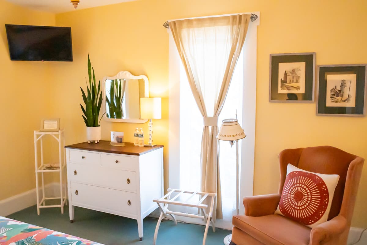 A cozy room featuring yellow walls, a window with sheer curtains, a dresser with a mirror, and a comfortable armchair.