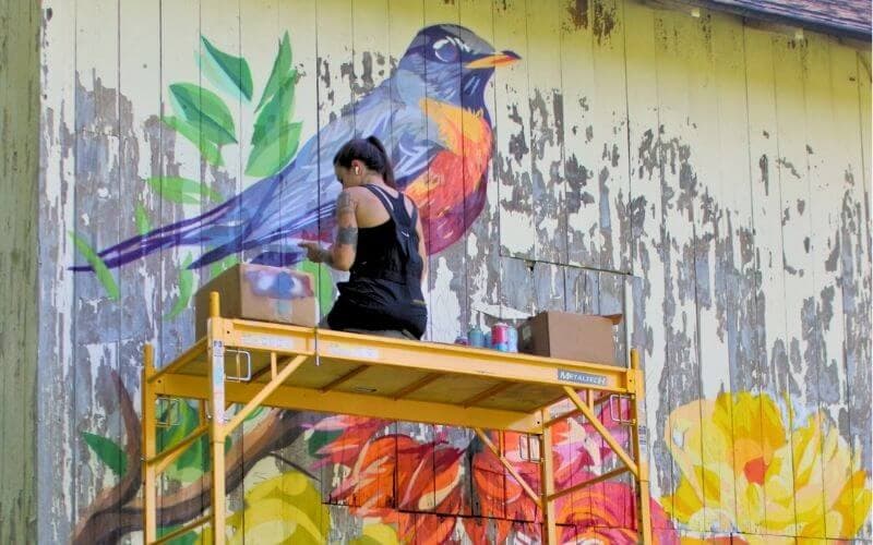 A person paints a colorful bird mural while working on a scaffold.