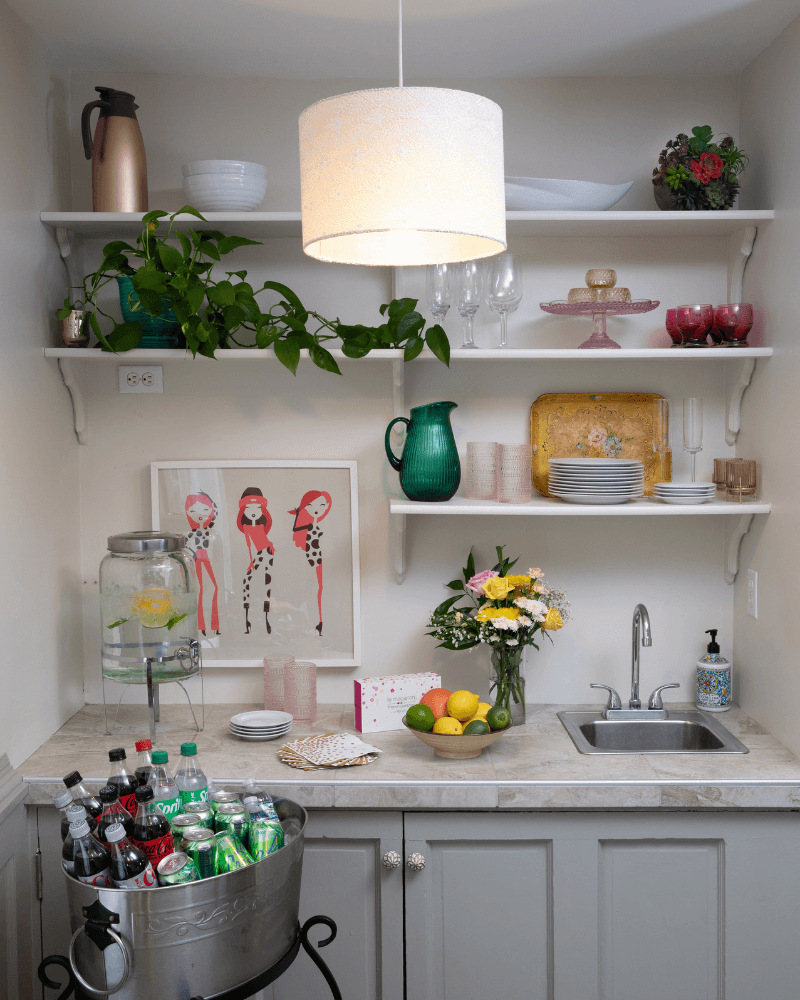A cozy kitchen corner featuring a water dispenser, colorful glassware, a sink, a flower arrangement, and a collection of beverages in a metal ice bucket.