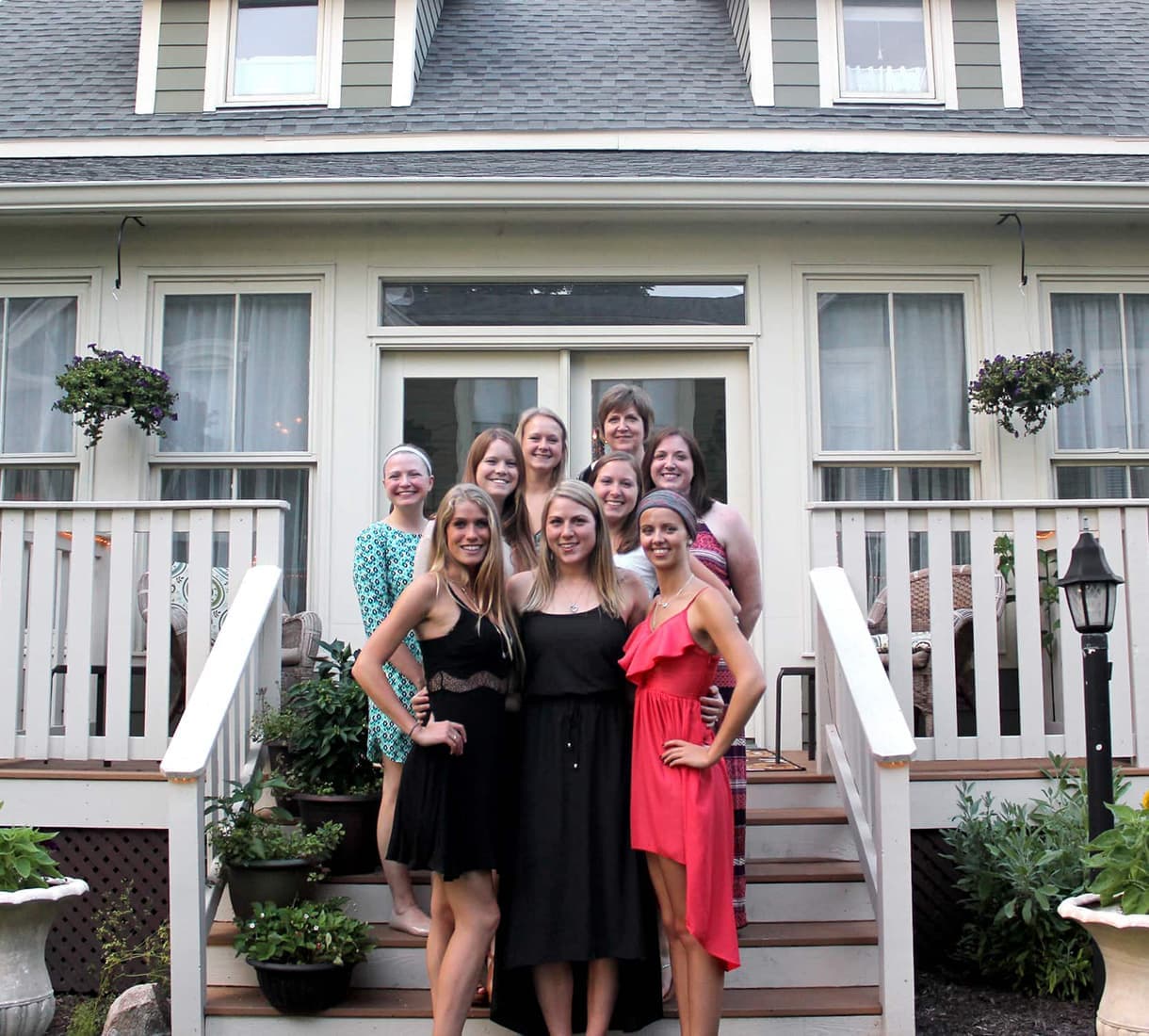 A group of ten women poses on the steps of a house, smiling and dressed in a variety of colorful outfits.