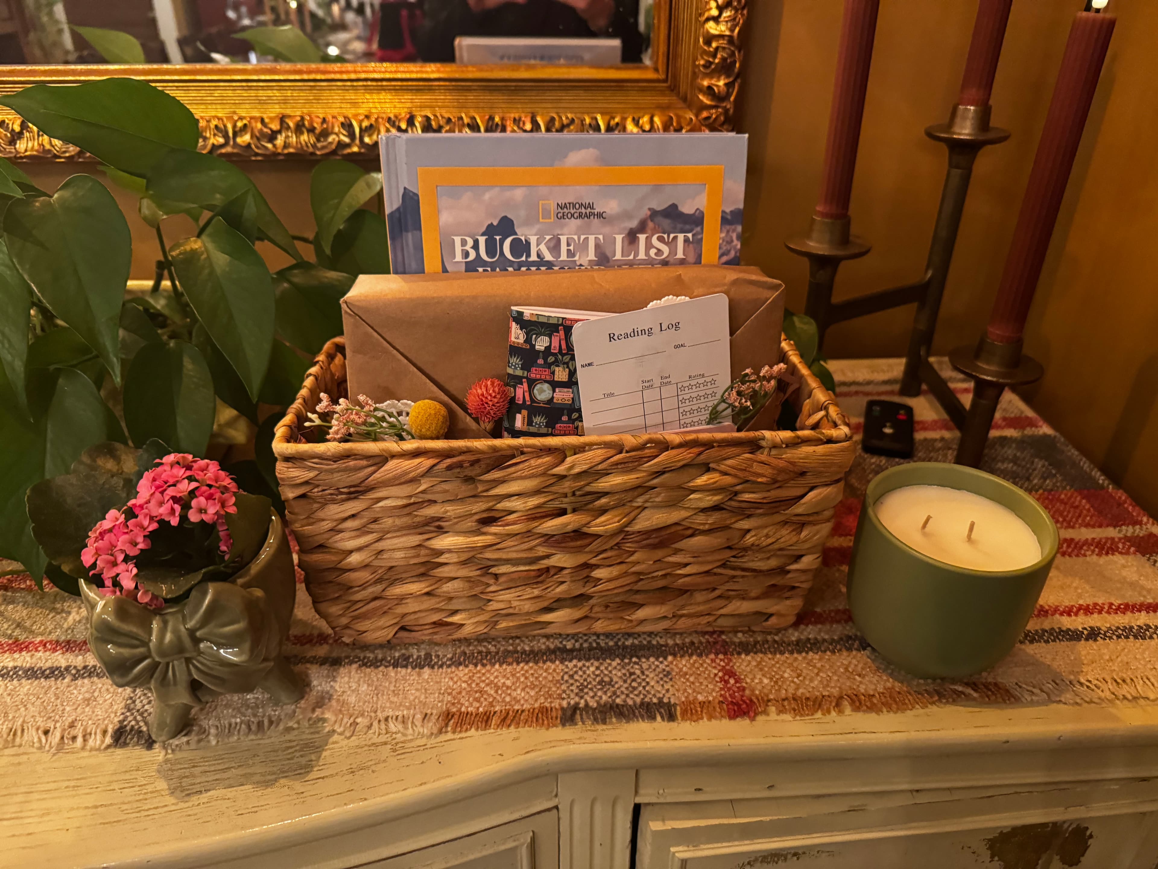 A woven basket filled with a "Bucket List" pamphlet, a brown envelope, a reading log, decorative items, and a candle on a patterned tablecloth.