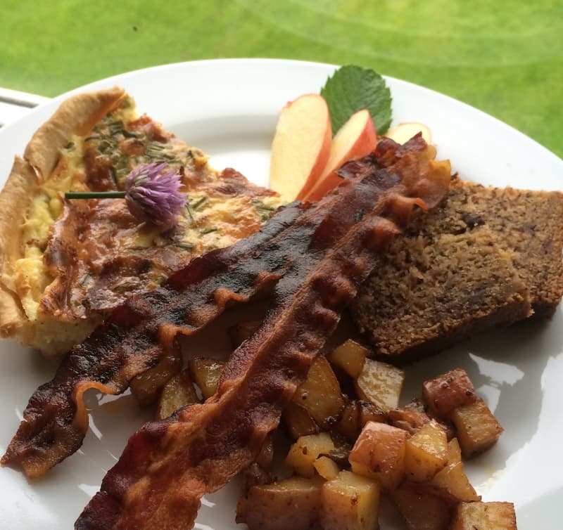 A plate of breakfast featuring quiche, crispy bacon, baked apples, a slice of bread, and a sprig of mint.