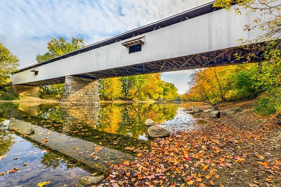 A covered bridge crosses a calm river surrounded by vibrant autumn foliage and fallen leaves.