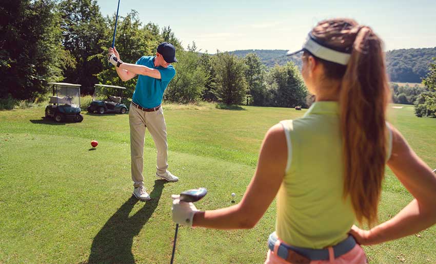 A golfer swings his club while a female player watches from a nearby tee. A golfer swings his club while a female player watches from a nearby tee.
