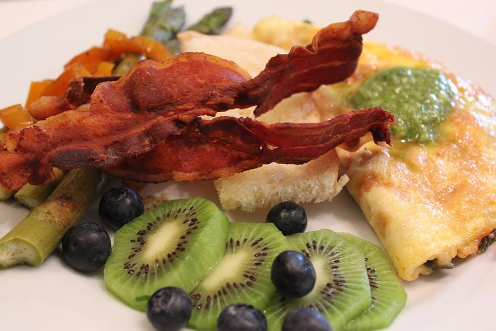 A plate featuring bacon, omelette, assorted vegetables, kiwi slices, and blueberries.