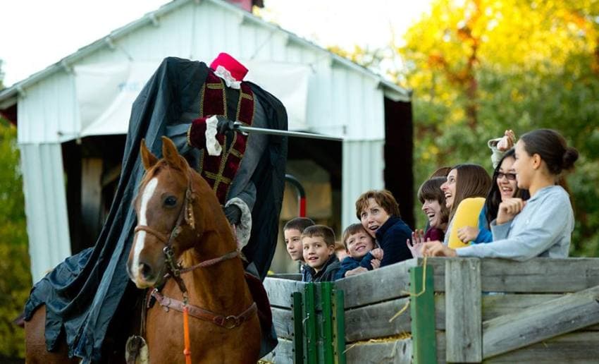 A headless horseman in a cape rides a horse while a group of excited children and adults watch from a wooden fence. A headless horseman in a cape rides a horse while a group of excited children and adults watch from a wooden fence.