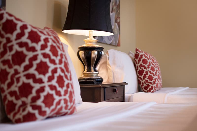 A cozy bedroom scene with two beds, red patterned pillows, and a lamp on a nightstand.