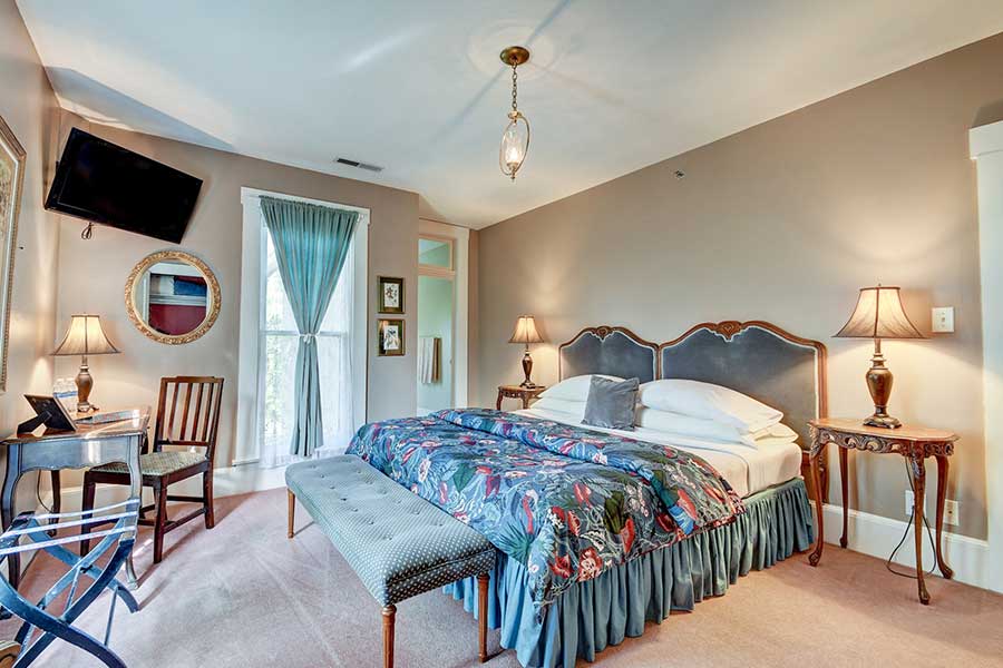 A cozy bedroom featuring a floral bedspread, bedside lamps, a chair, and large windows with sheer curtains.