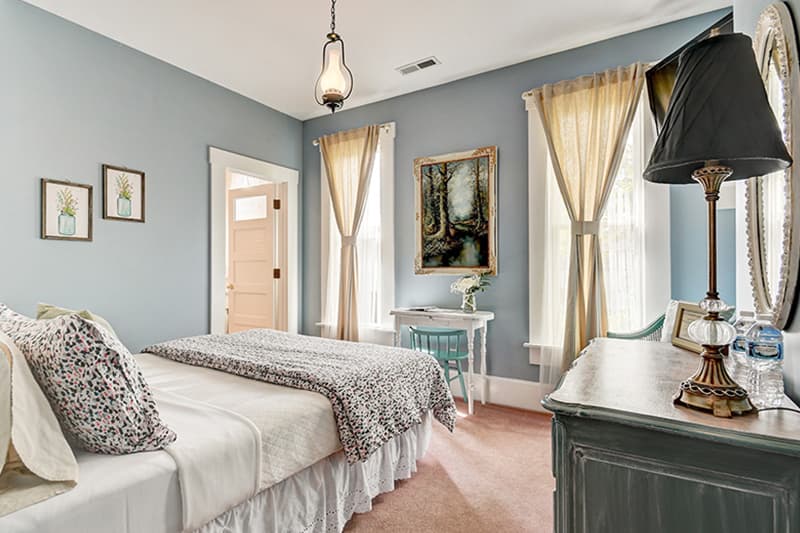 A cozy bedroom featuring light blue walls, two beds with decorative linens, large windows with sheer curtains, and vintage furnishings.