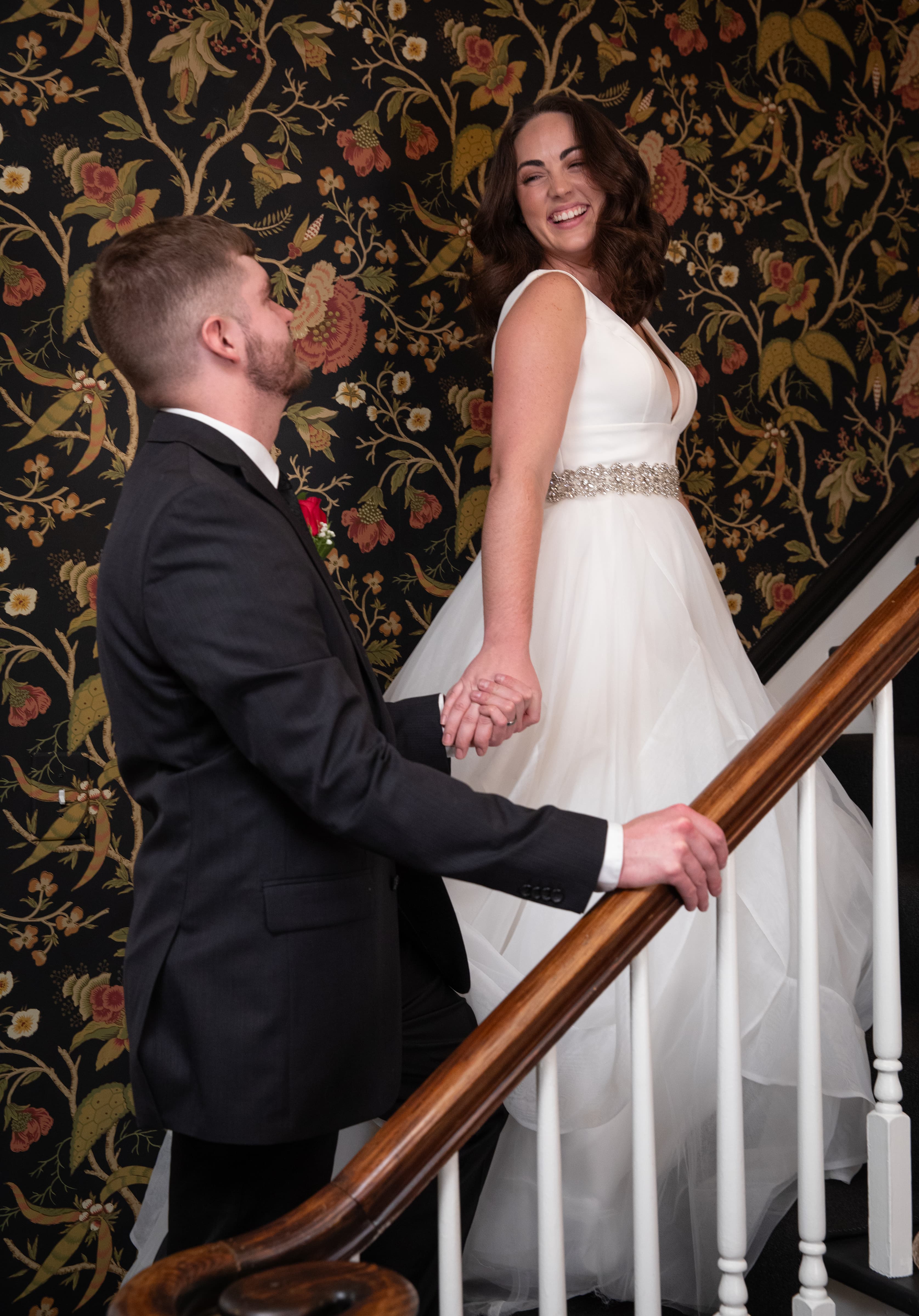 A couple holds hands on a staircase, smiling at each other against a floral wallpaper backdrop.