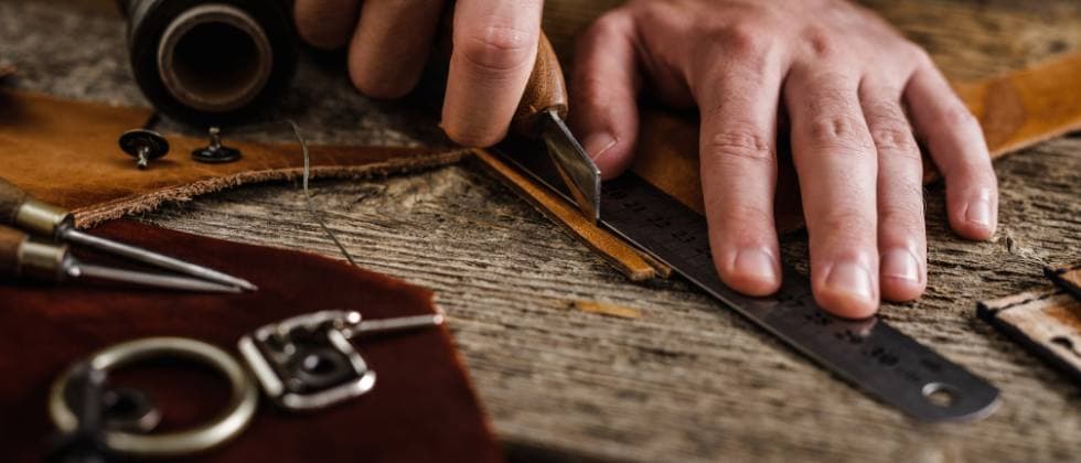 A hand uses a knife to cut leather on a wooden table, surrounded by tools and materials. A hand uses a knife to cut leather on a wooden table, surrounded by tools and materials.