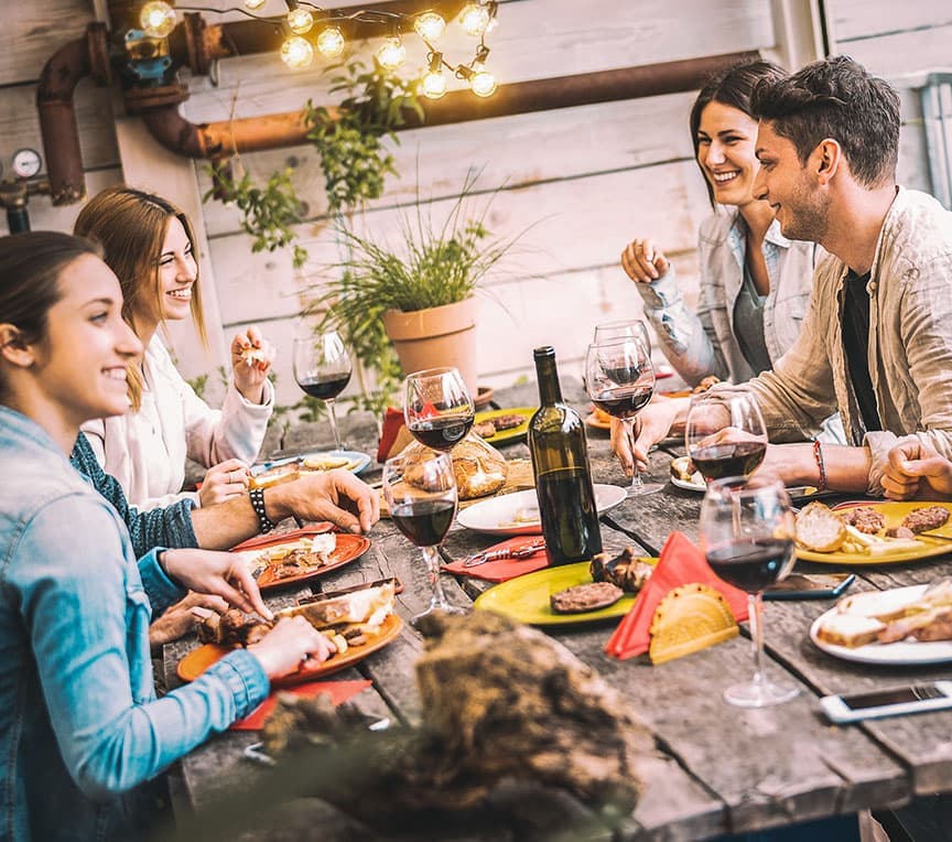 A joyful group of friends enjoys a meal outdoors, surrounded by wine glasses and colorful plates.