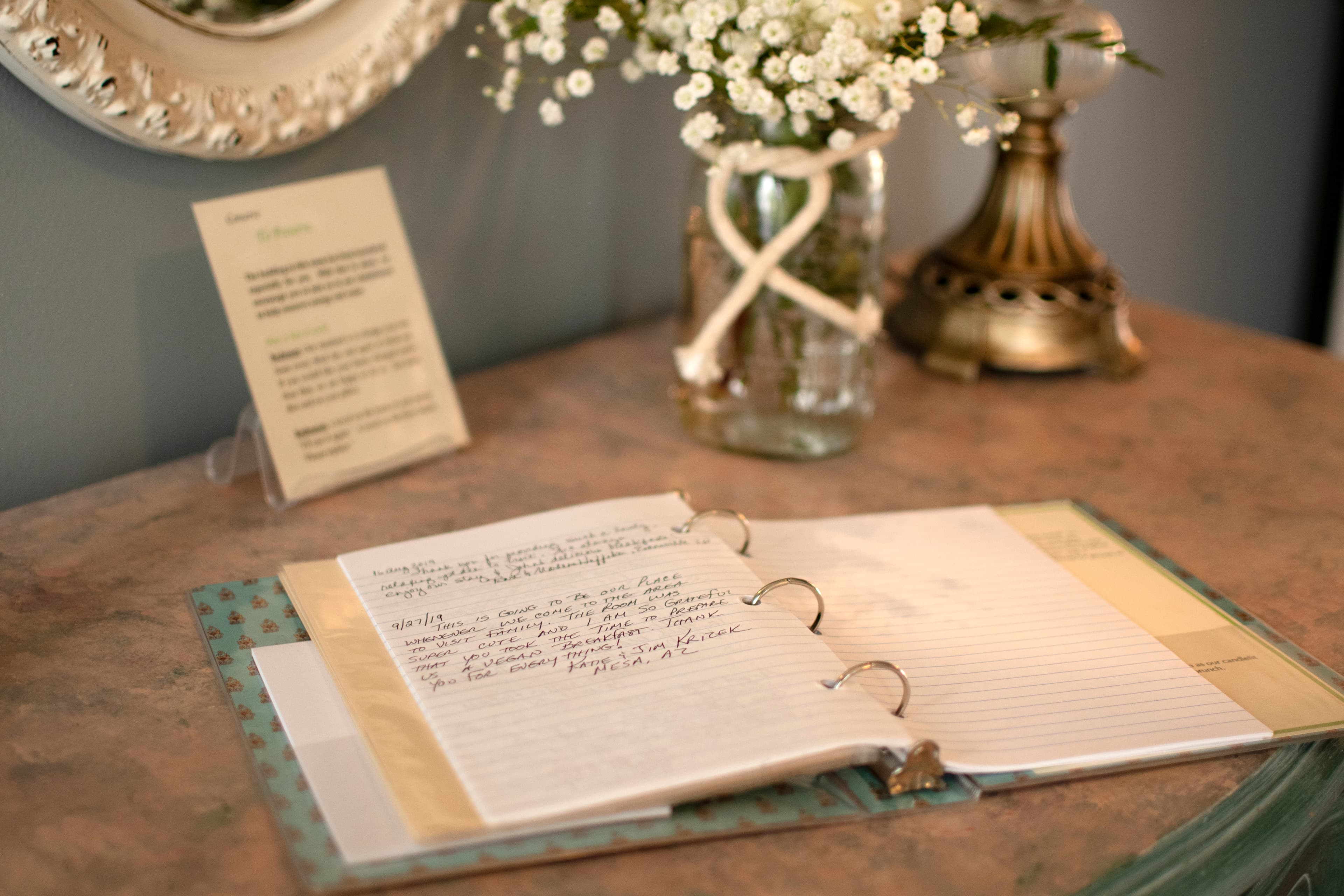 An open binder with handwritten notes sits on a table beside a vase of flowers and a display card.