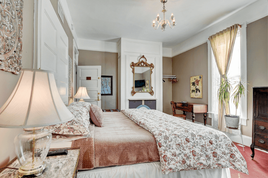 A cozy bedroom featuring floral-patterned bedding, a vintage lamp, and a large window with drapes.